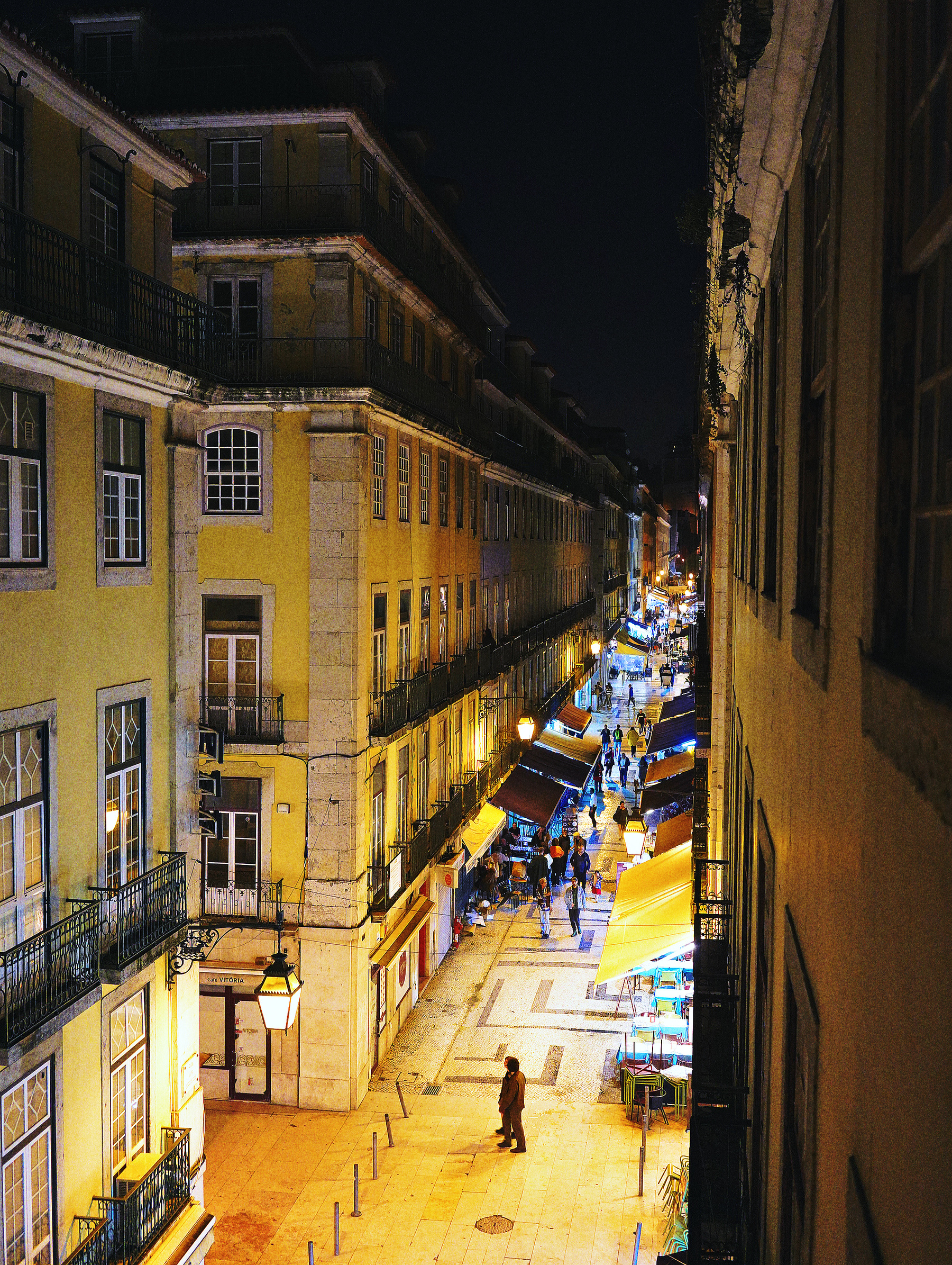 A vibrant, narrow street in a European city is bustling with people and illuminated by warm streetlights and colorful market stalls at night.