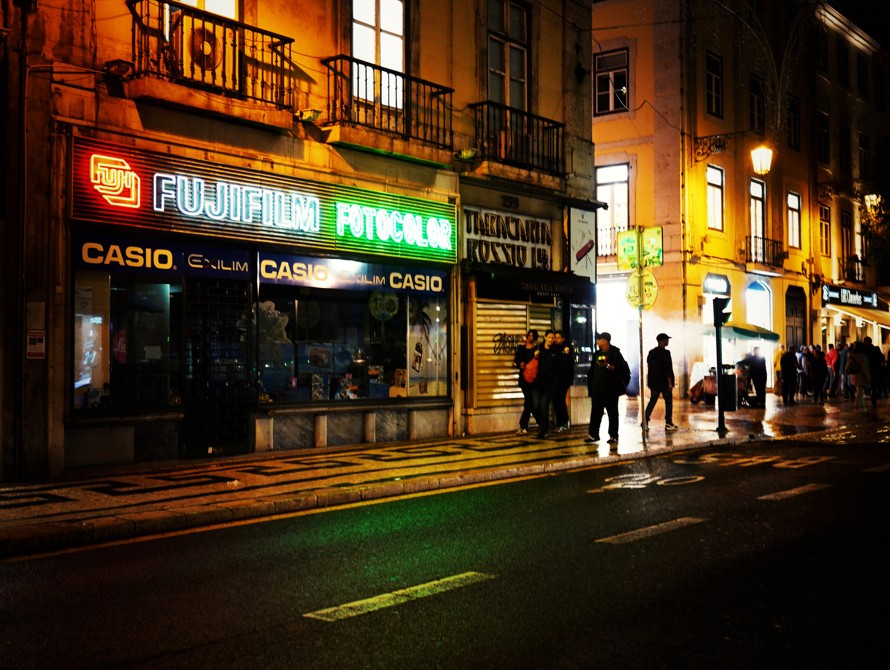 A city street at night is illuminated by neon signs advertising brands like Fujifilm and Casio, with people walking along the sidewalk.