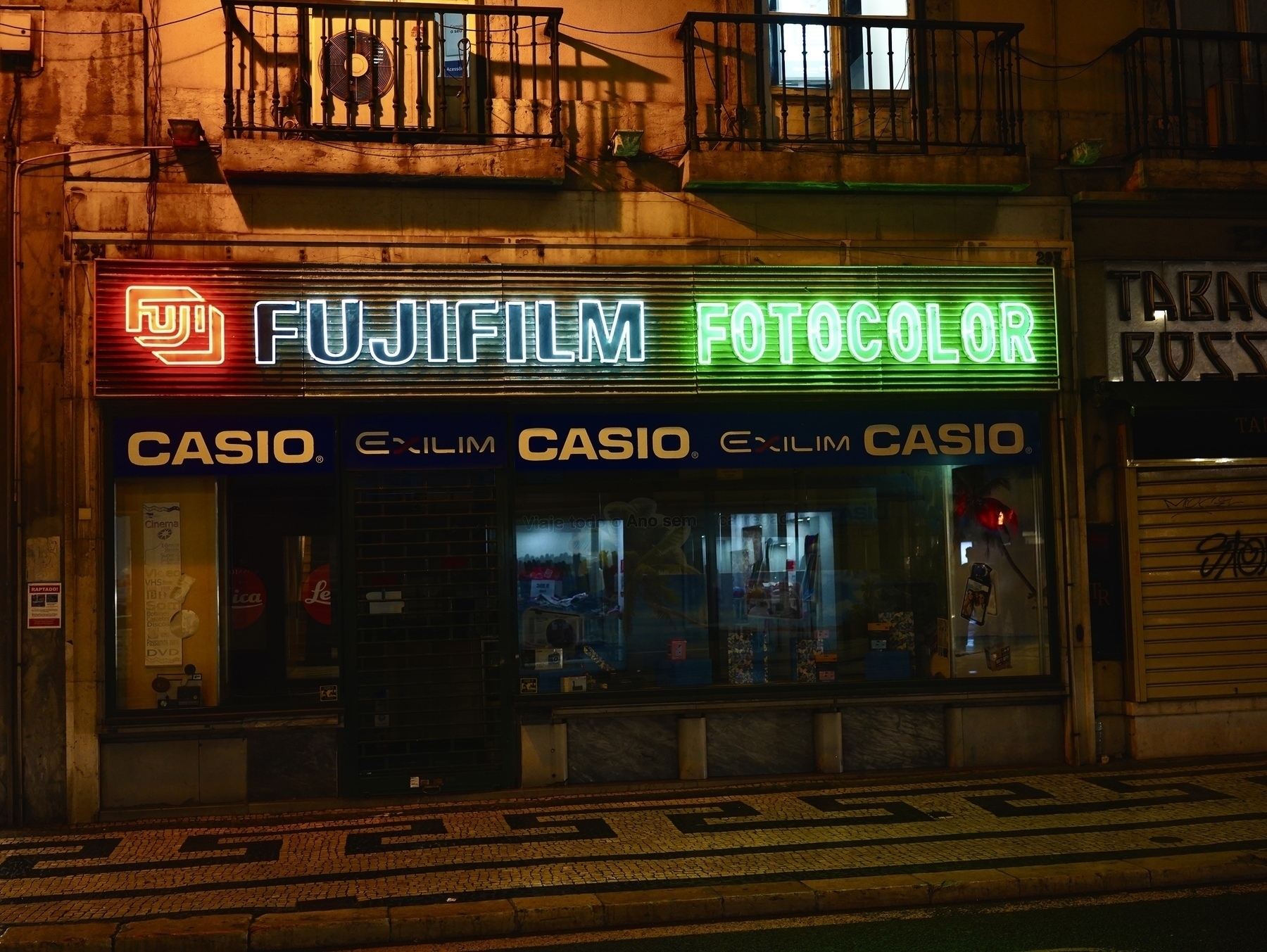 A storefront displays neon signs for Fujifilm and Casio on a dimly lit street.