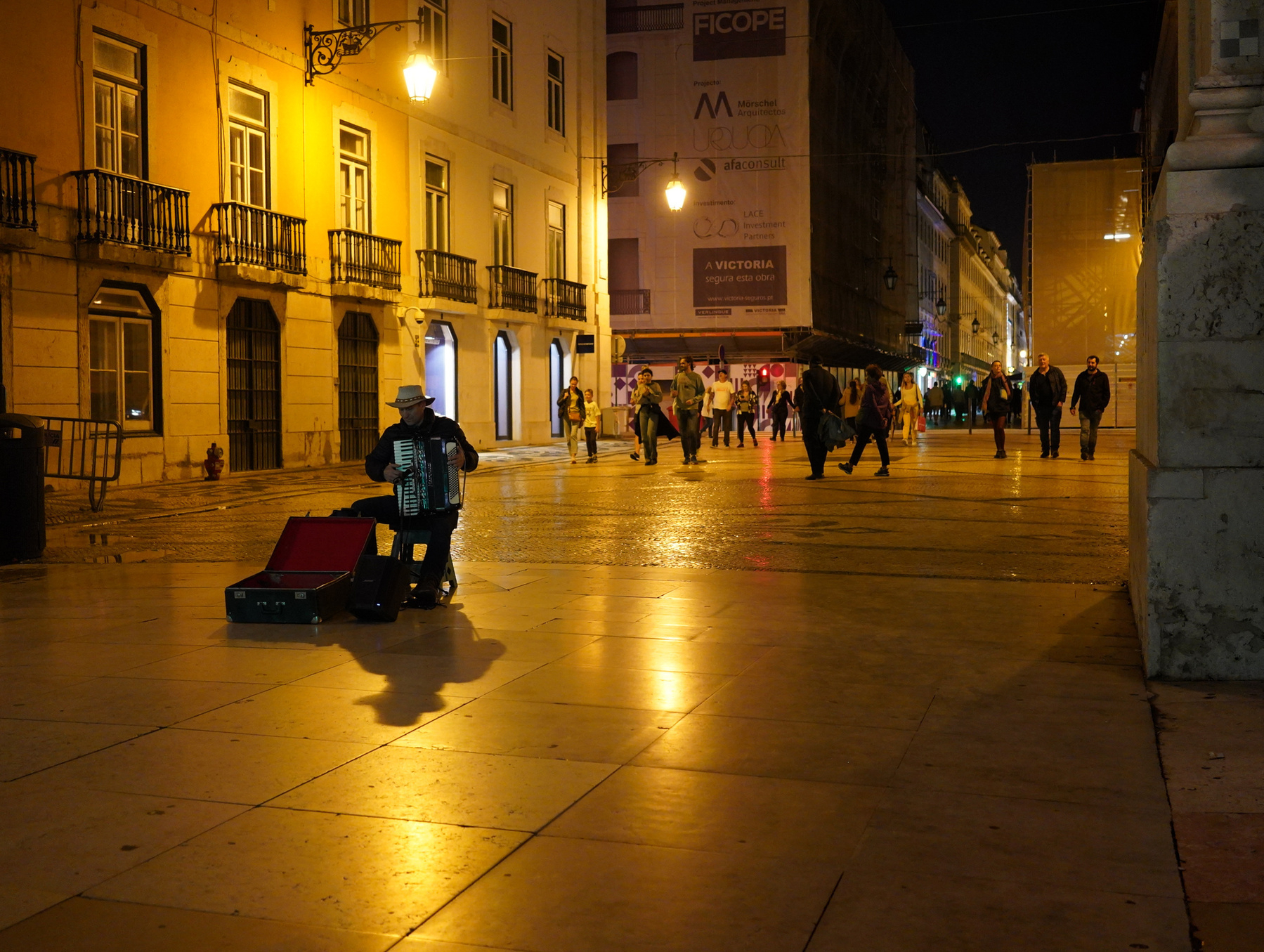 A street musician plays the accordion under a streetlamp on a cobblestone path as people walk by at night in a city setting.