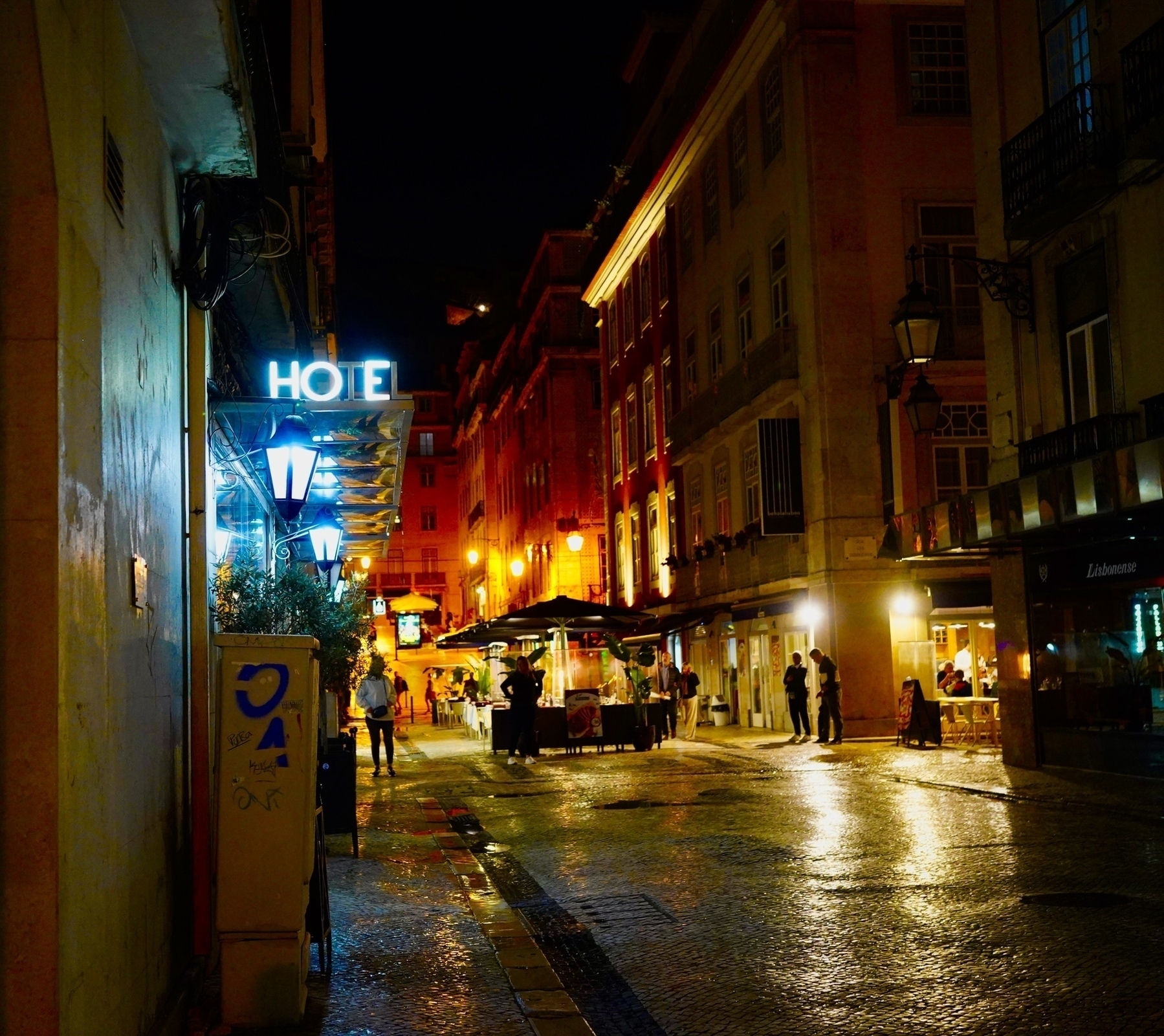 A dimly lit street scene at night features a HOTEL sign, illuminated buildings, and people walking along the wet cobblestone path.