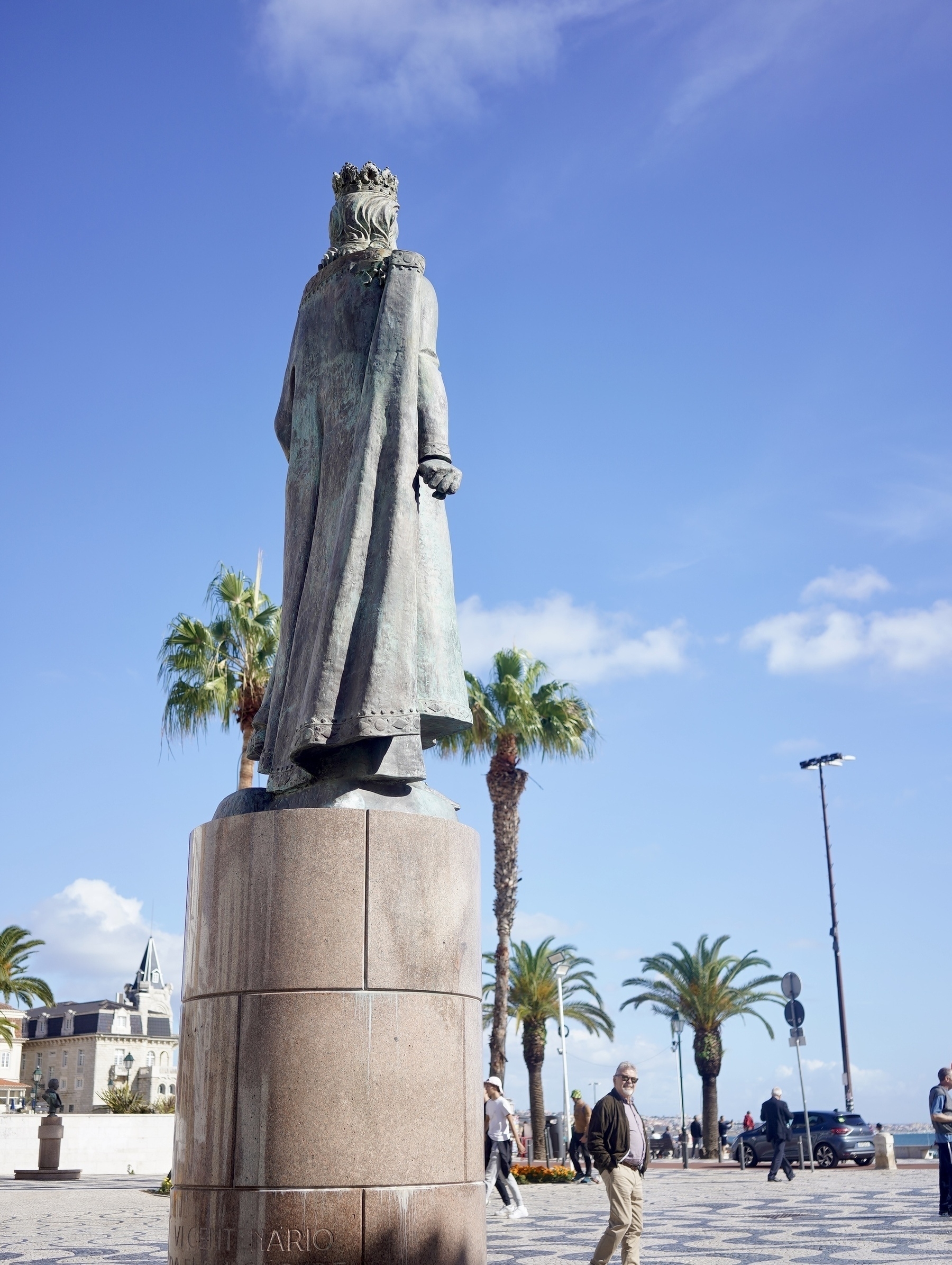 A statue of a king wearing a crown stands atop a pedestal with palm trees and people in the background.