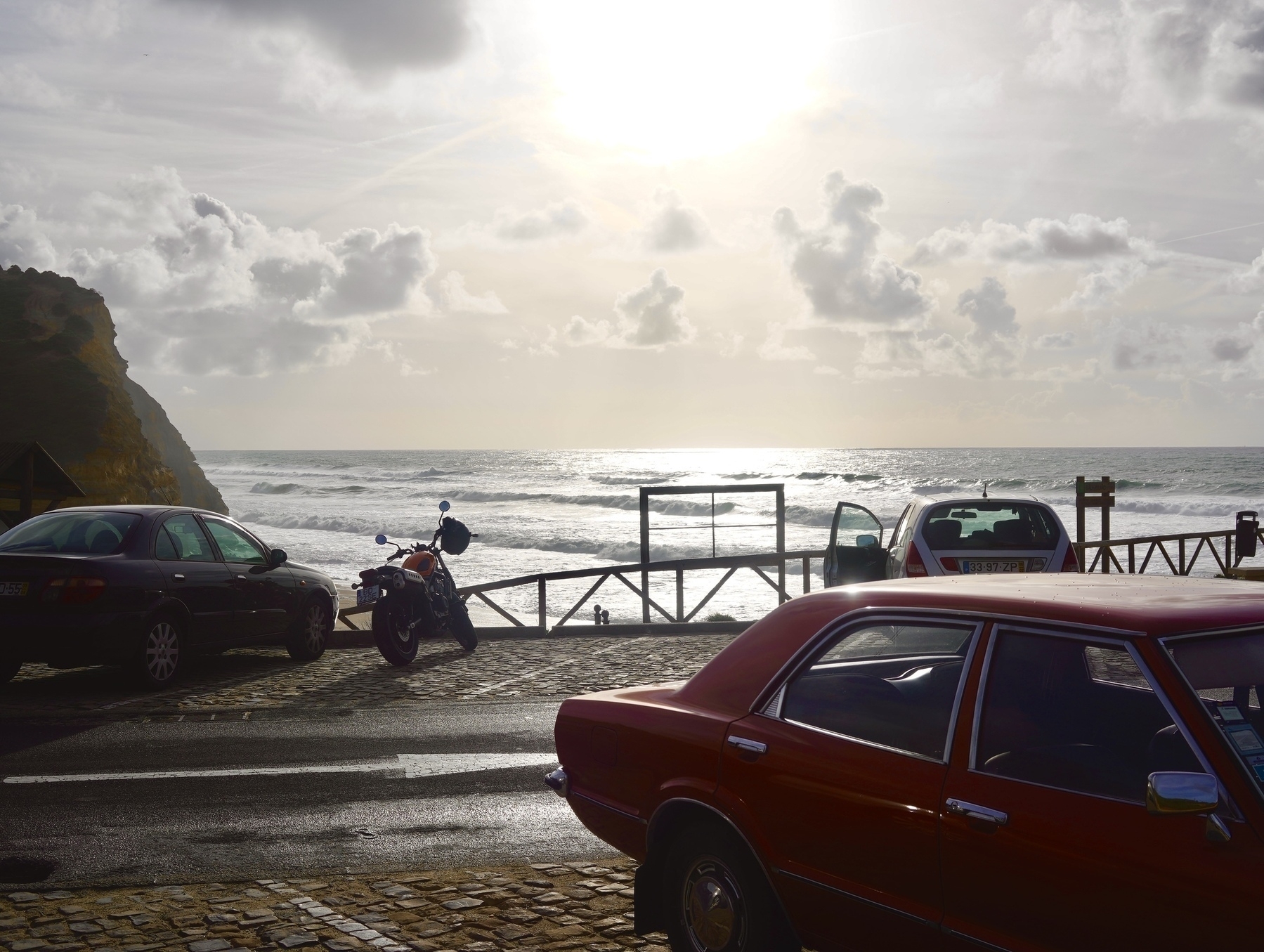 A coastal scene at sunset features parked cars, a motorcycle, and a person near a railing overlooking the ocean.