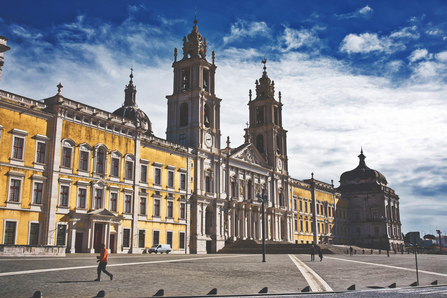 A grand, historic building with intricate architecture stands against a partly cloudy sky, with a person and a car visible on the cobblestone path in front.