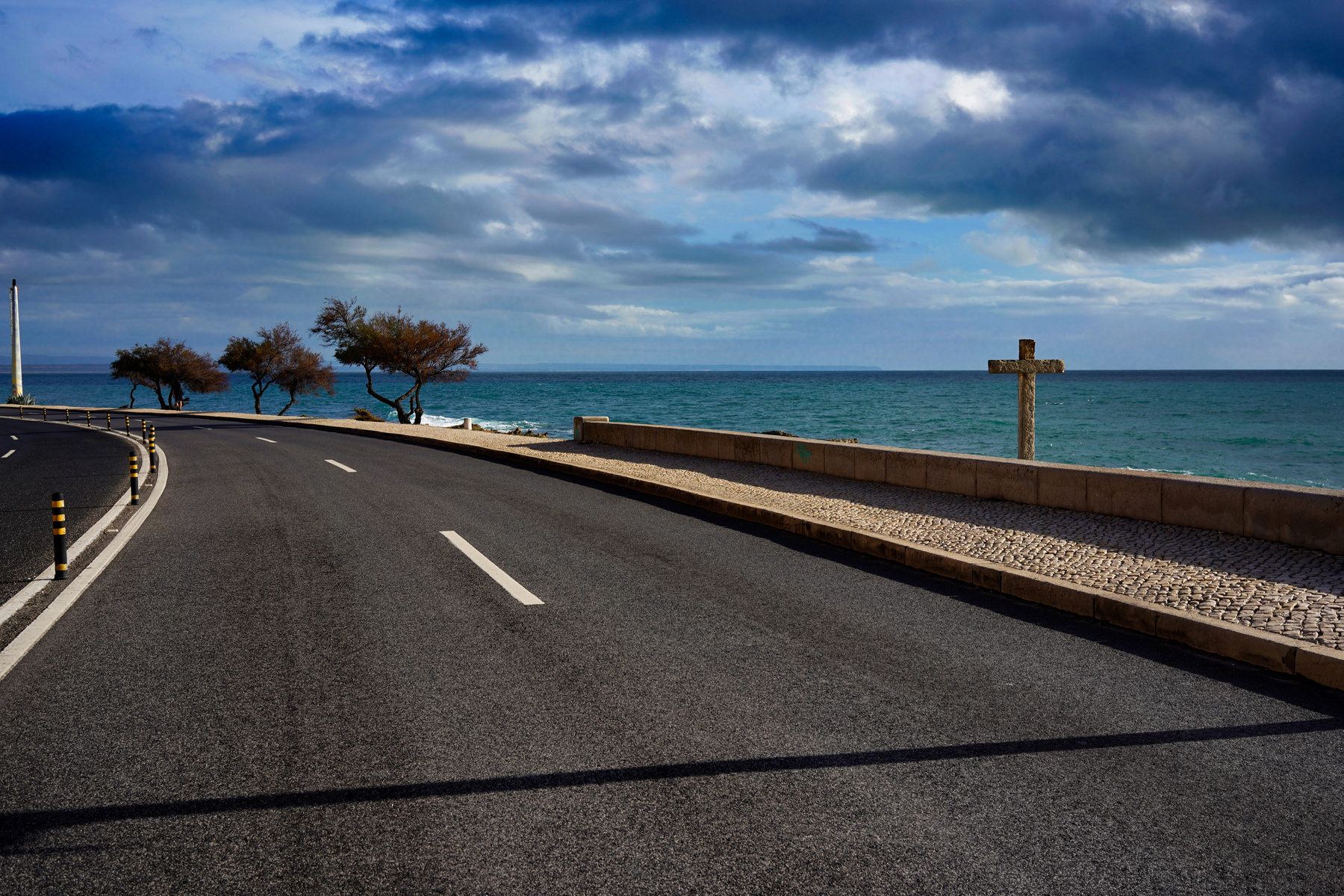 A coastal road curves alongside the ocean as clouds gather above, with a cross visible on the side.