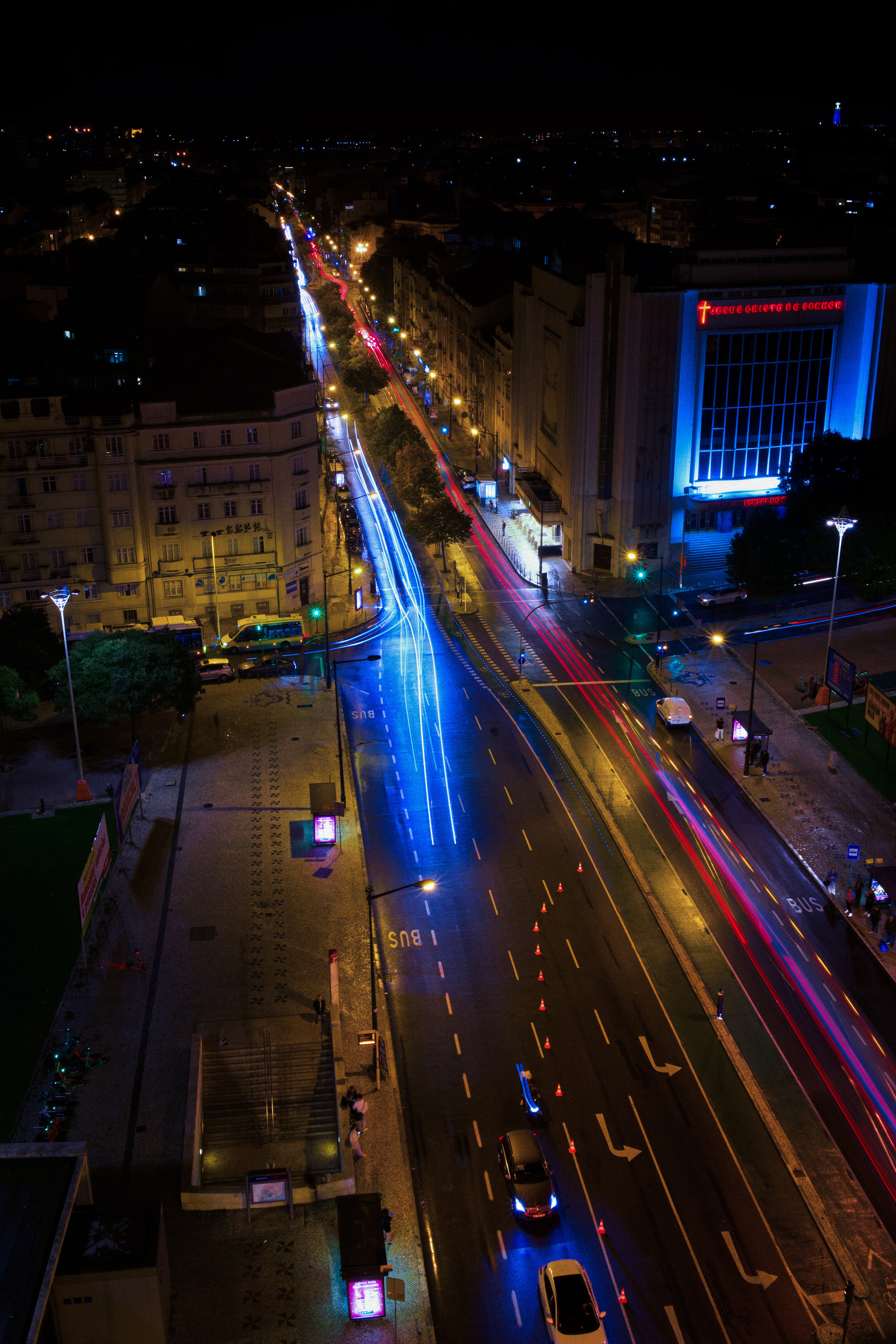 A city street at night is illuminated by blue and red light trails from moving vehicles, with buildings and streetlights lining the road.