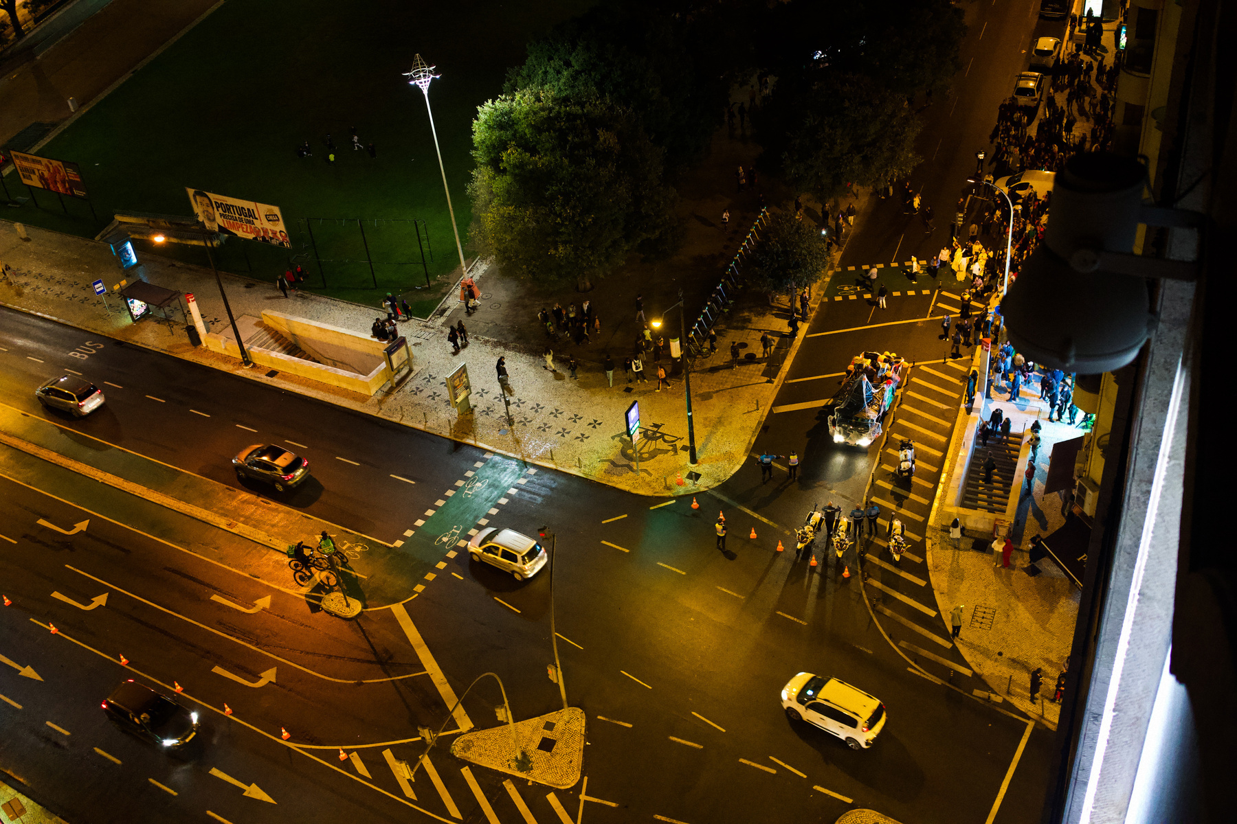 Aerial view of a lively urban street at night with cars, pedestrian activity, and a well-lit gathering near a stairway.