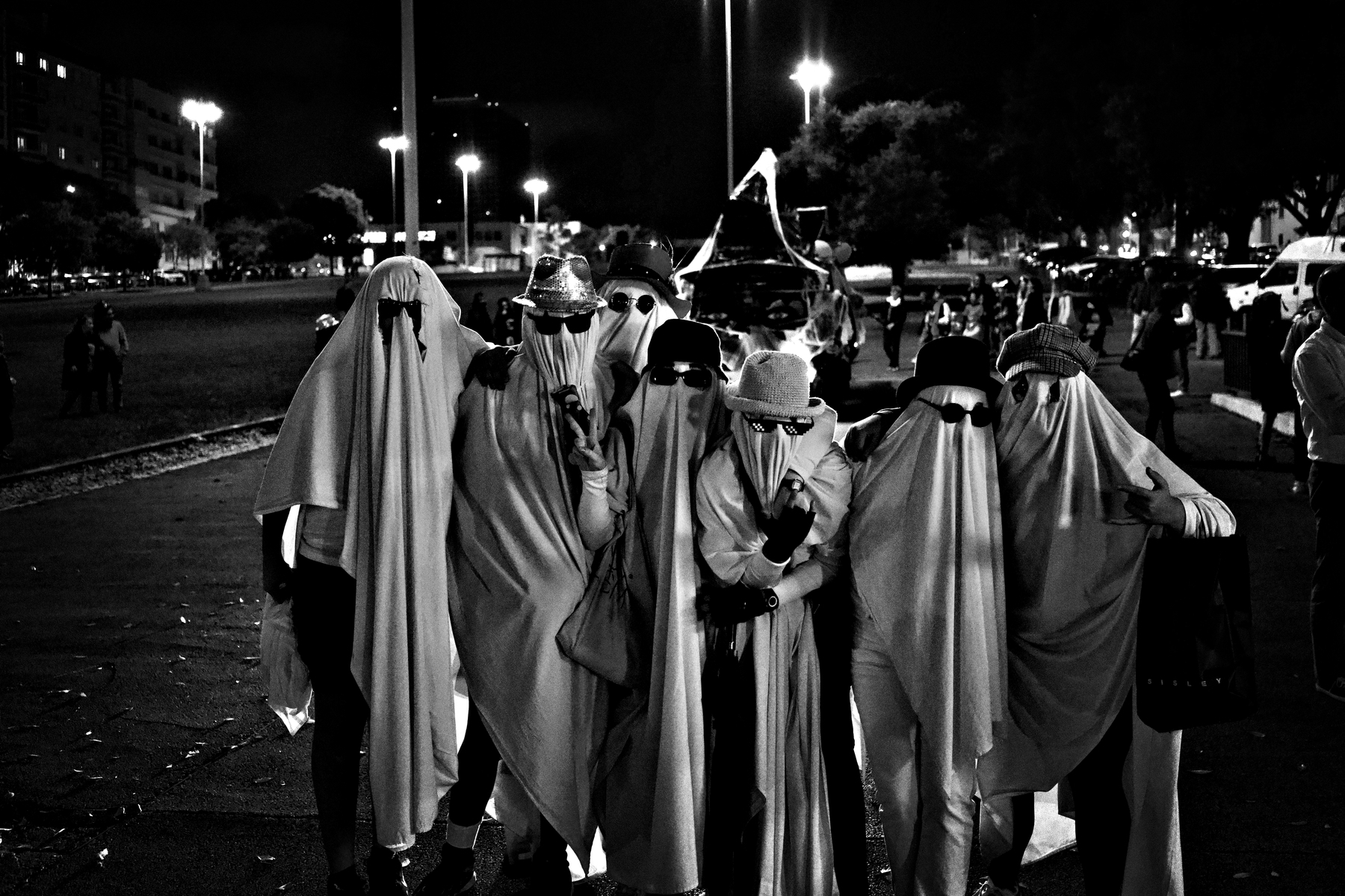 A group of people dressed as ghosts, wearing sheets and sunglasses, pose together at night in an urban setting.