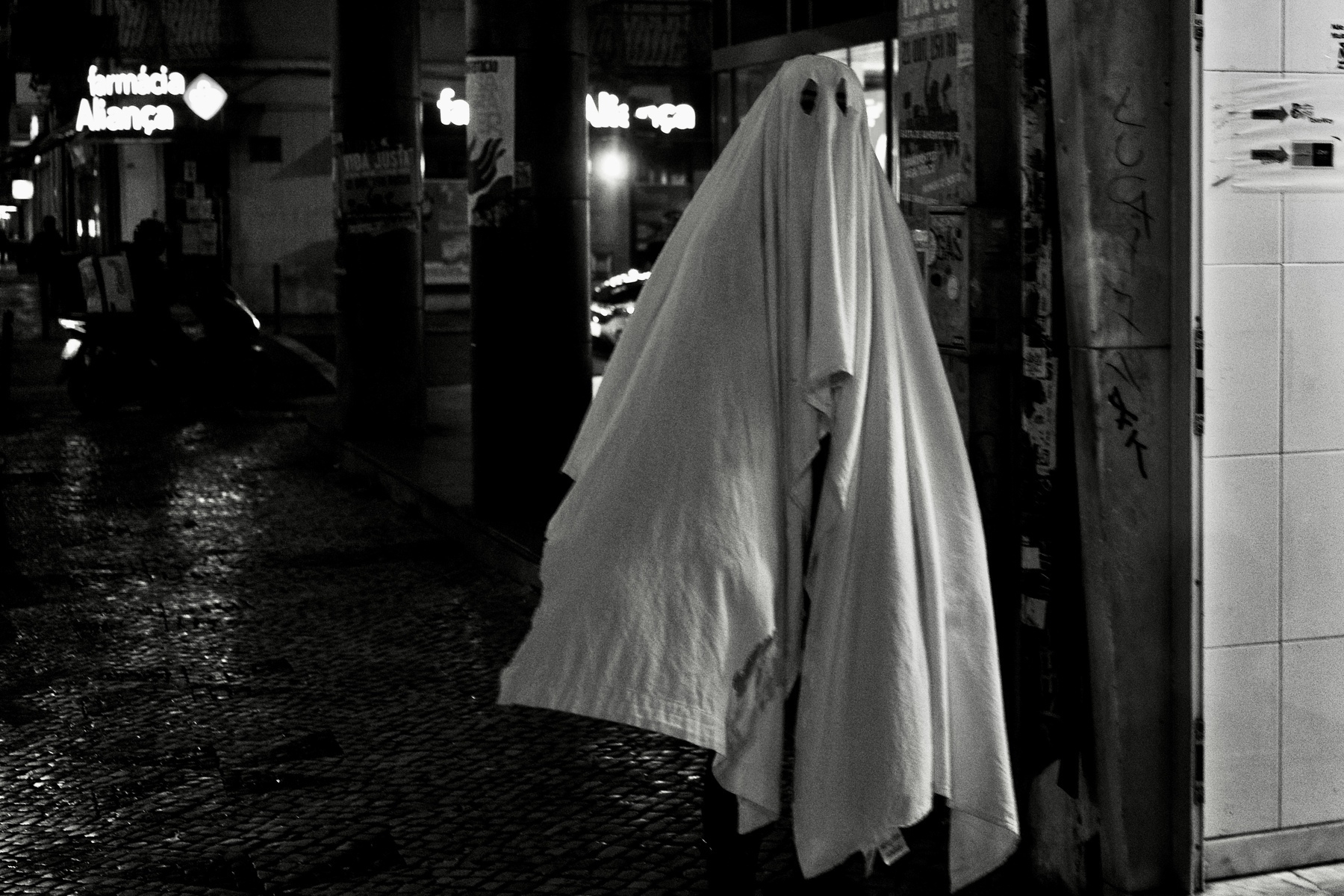 A person is dressed as a traditional ghost with a white sheet in an urban nighttime setting.