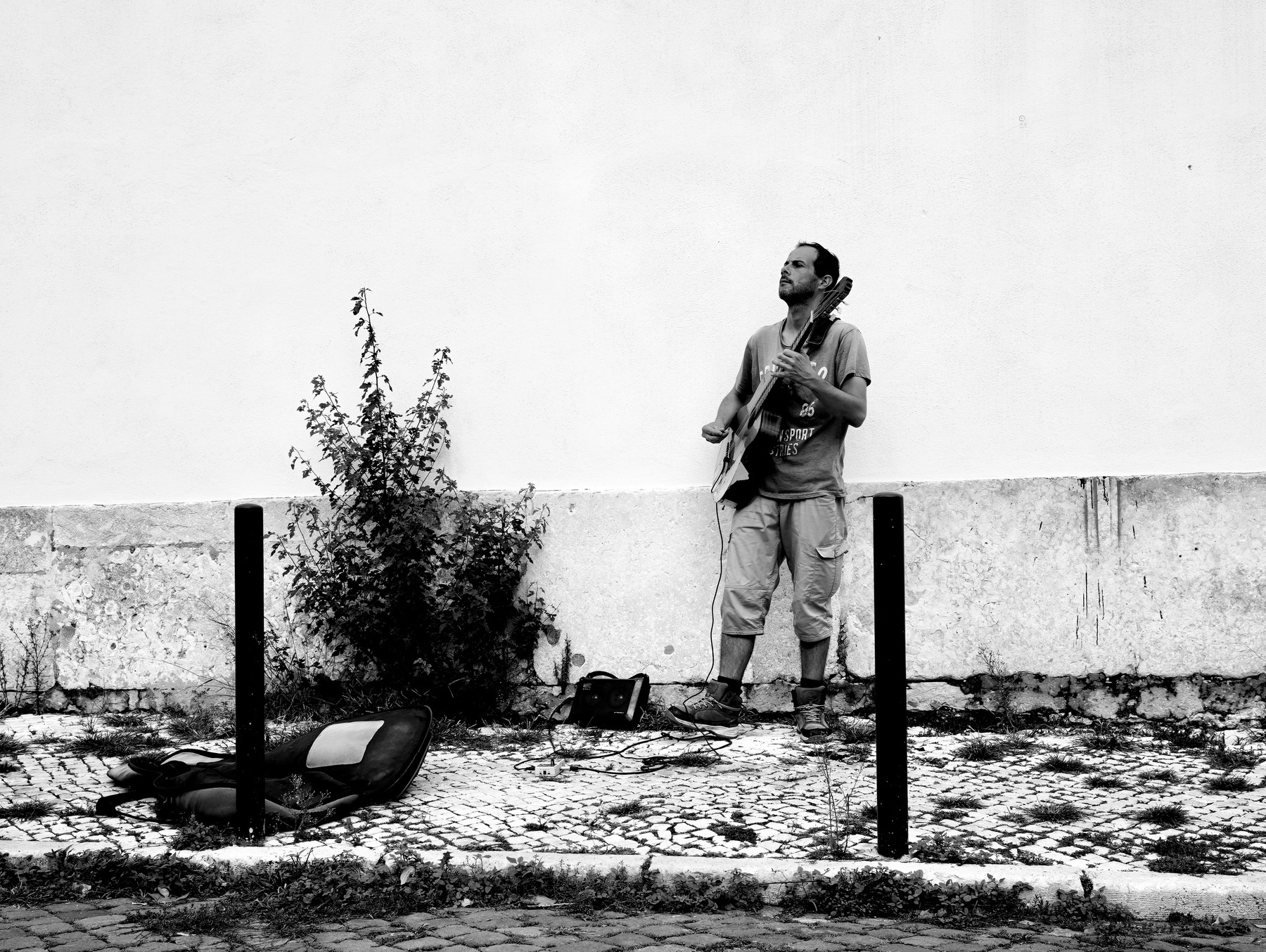 A man stands on a cobblestone path playing a guitar next to a wall, with an open guitar case on the ground in front of him.