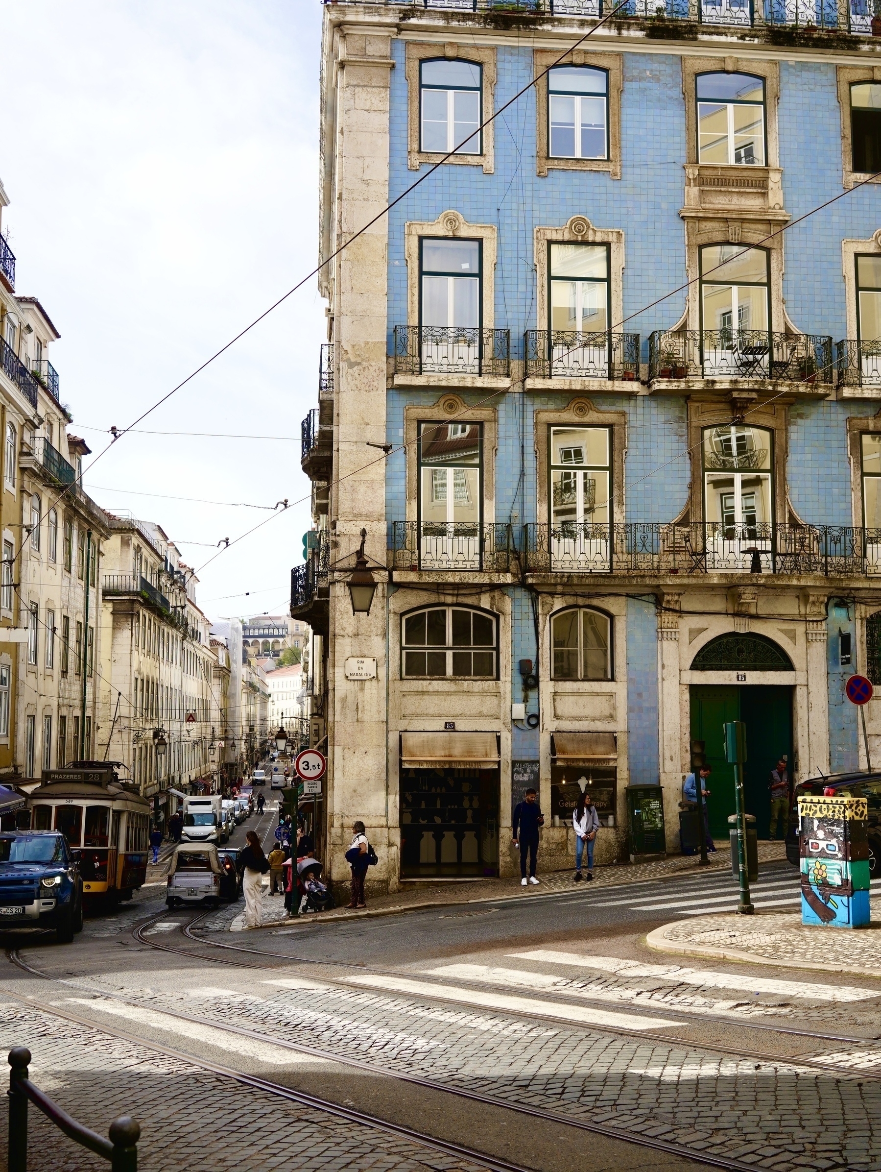 A street scene in a European city features a corner building with blue tiles, tram lines, cobblestone roads, and pedestrians.