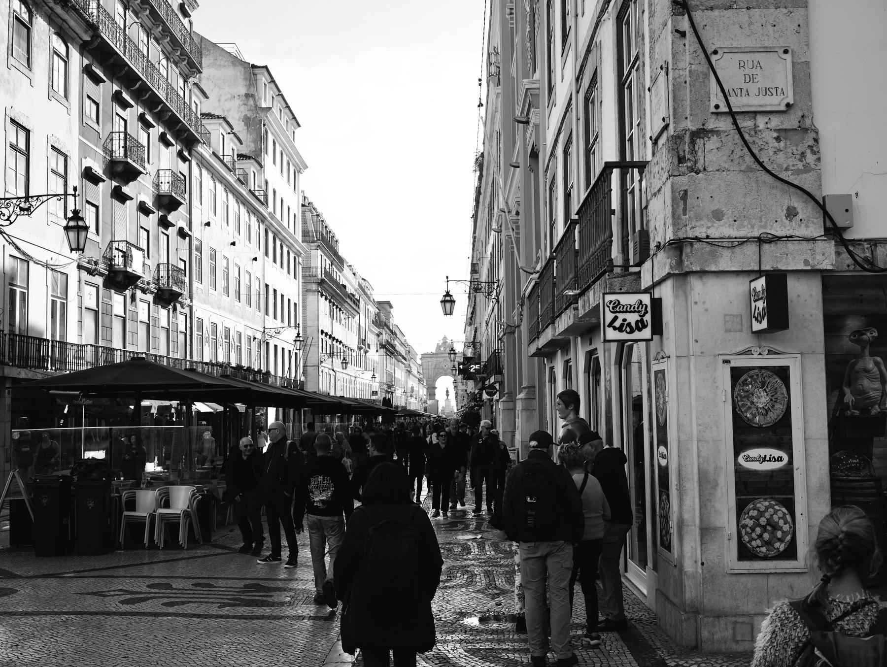 A busy street scene in black and white features people walking along a cobblestone path lined with buildings and outdoor cafés.