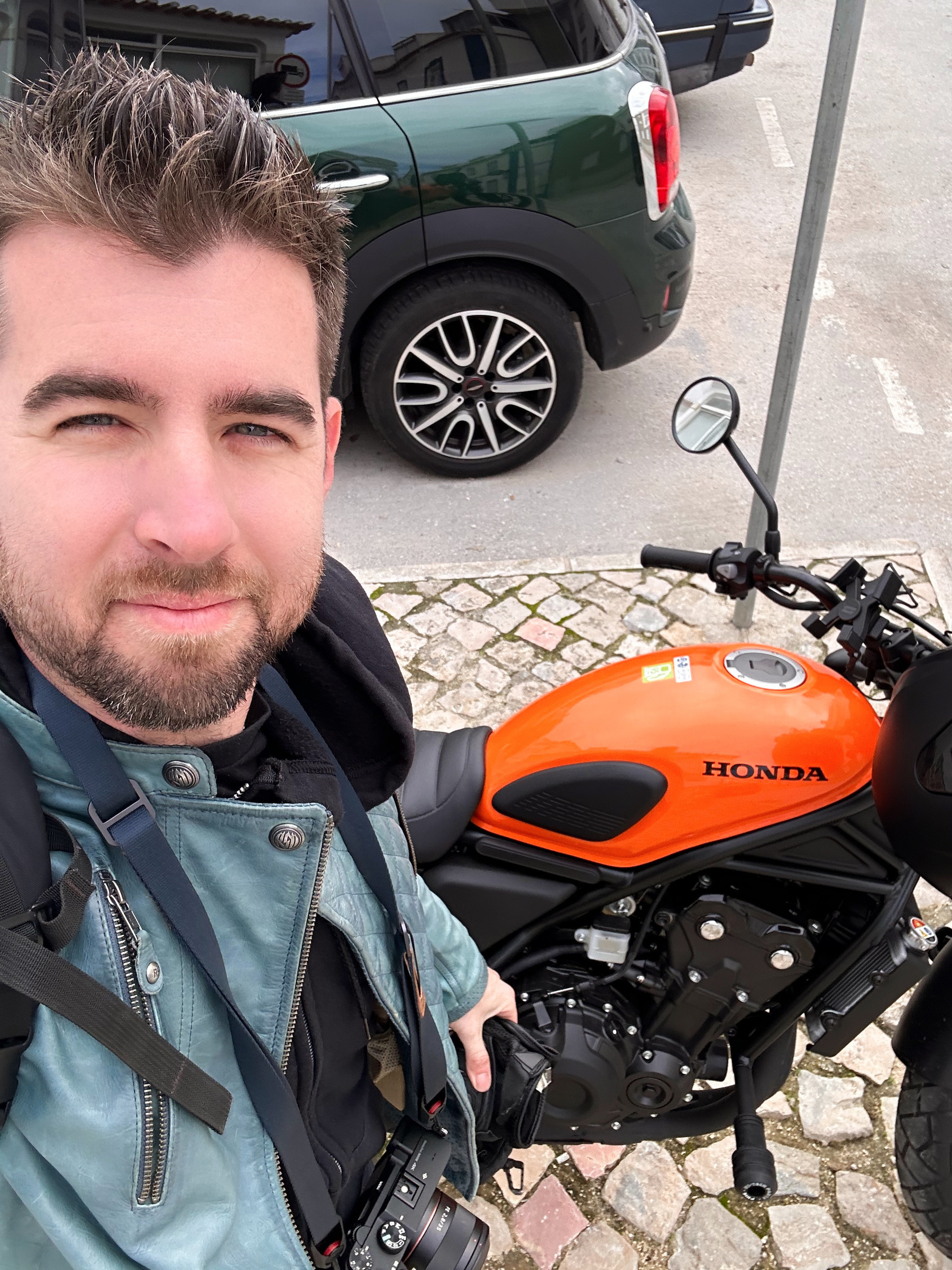 A person is taking a selfie next to an orange Honda motorcycle parked beside a green car.