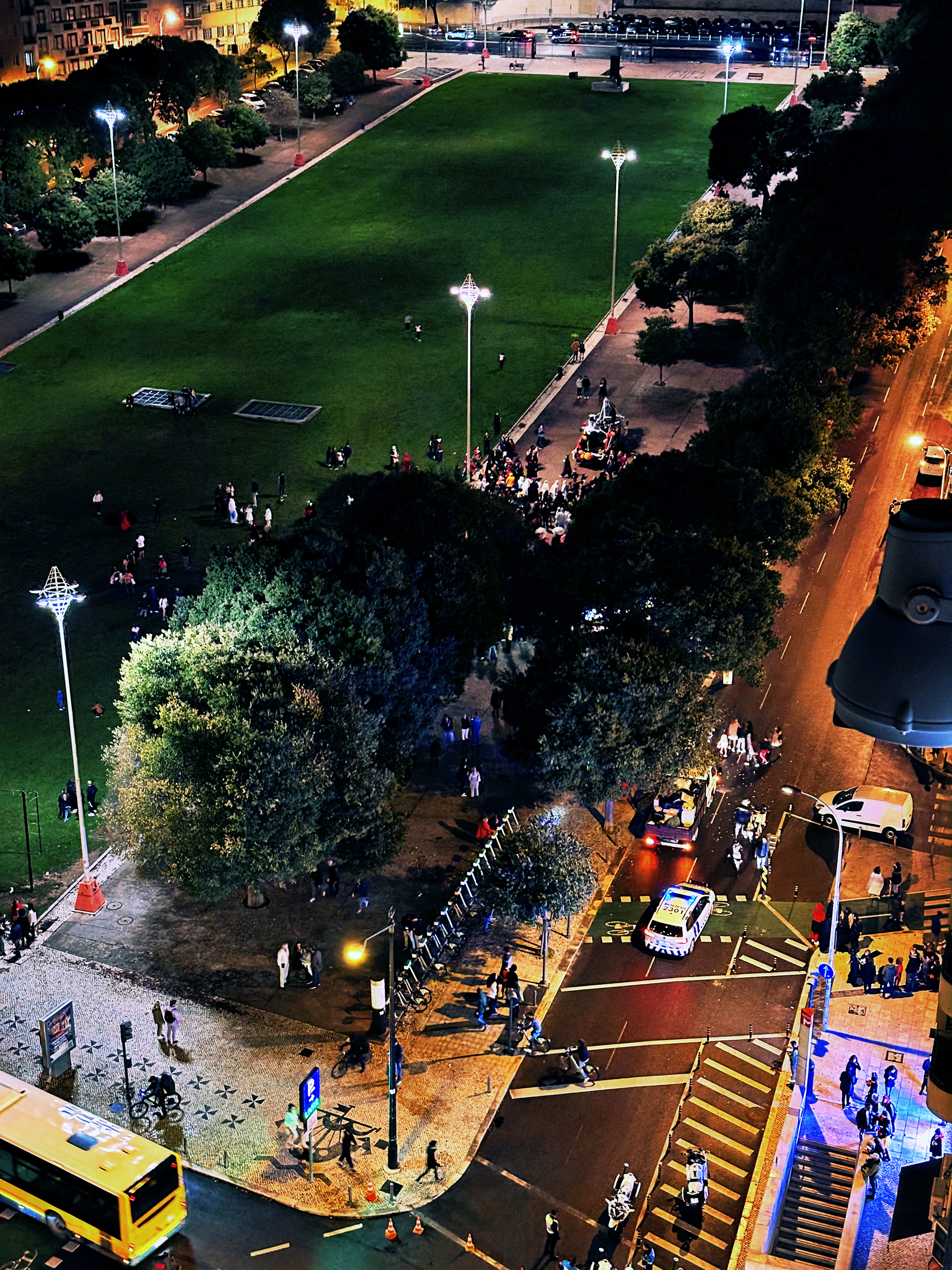 A vibrant city scene at night features a park, trees, pedestrians, streetlights, and a bus.